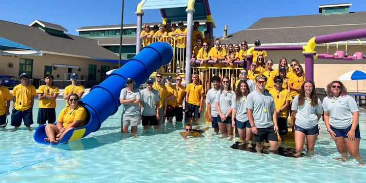 A group of lifeguards and staff in yellow shirts poses by a pool with a blue slide on a sunny day.