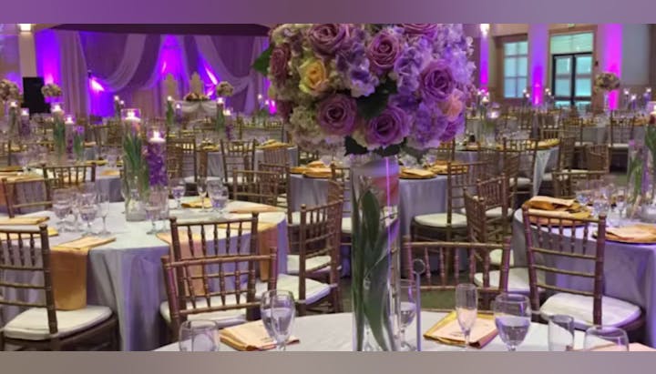 Elegant event setup with purple lighting, floral centerpieces, round tables with tableware, and golden chairs.