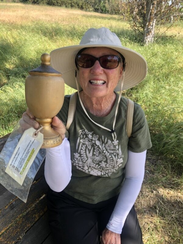 A smiling woman wearing a sun hat and sunglasses, holding an urn with a "Golden" tag, sitting outdoors.