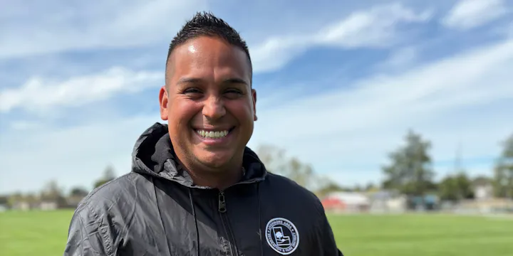 A man smiles broadly outdoors, wearing a jacket with a logo, standing on a green field under a blue sky.