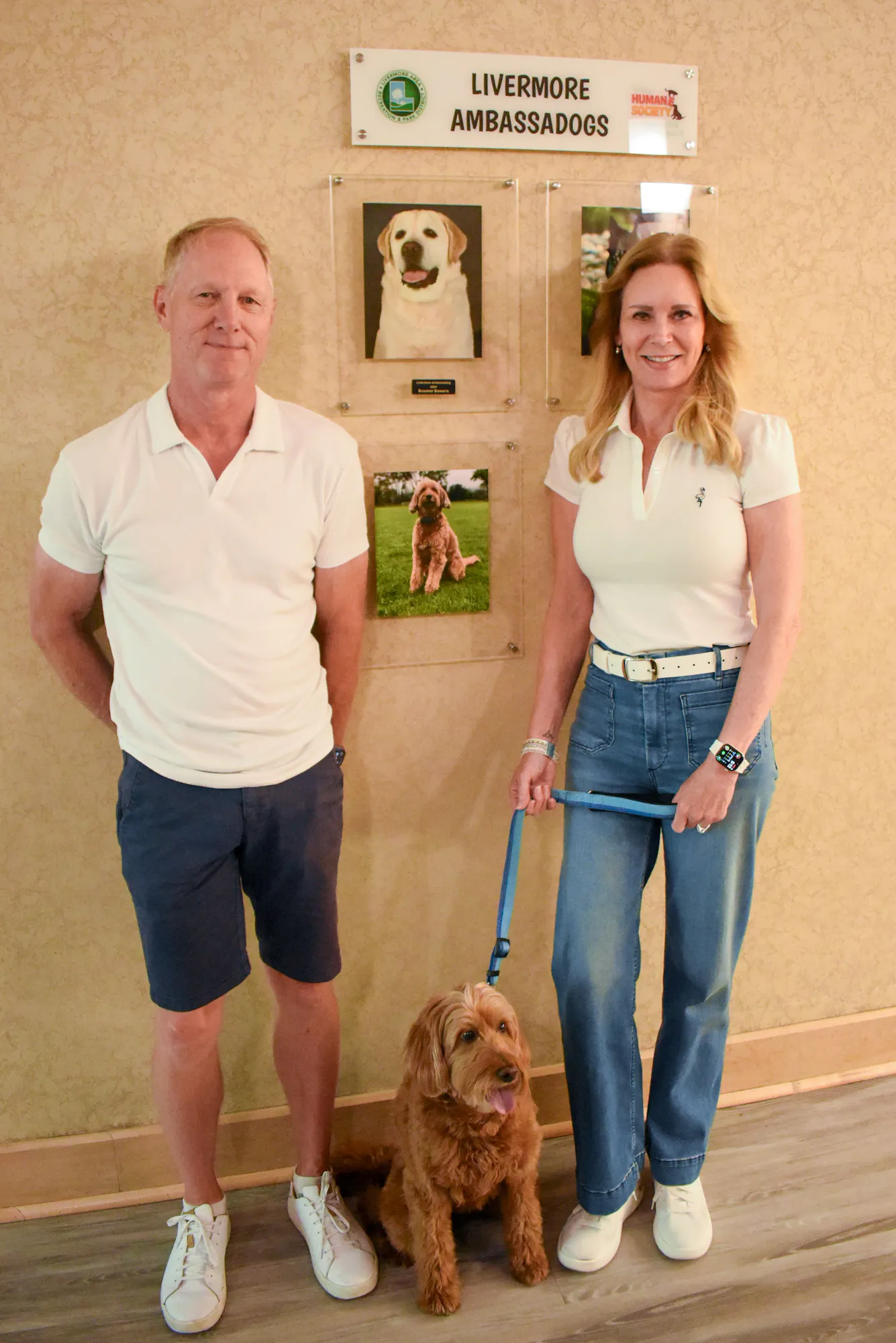 A man and a woman pose with a dog in front of a "Livermore Ambassadogs" display showcasing dog photos.