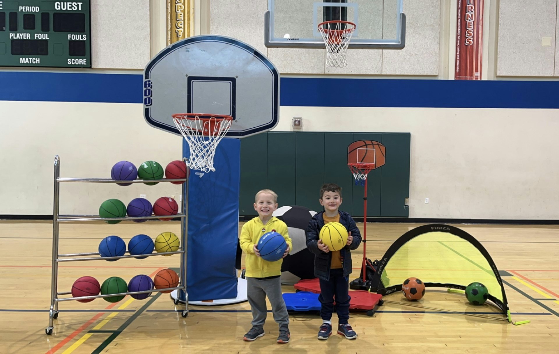 Two children in a gym with basketballs, hoops, a rack of balls, and soccer goals.