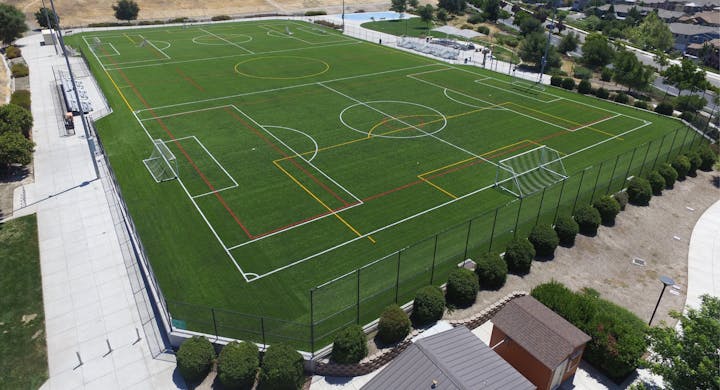 An aerial view of a multisport artificial turf field with soccer and other sports lines, surrounded by a fence.