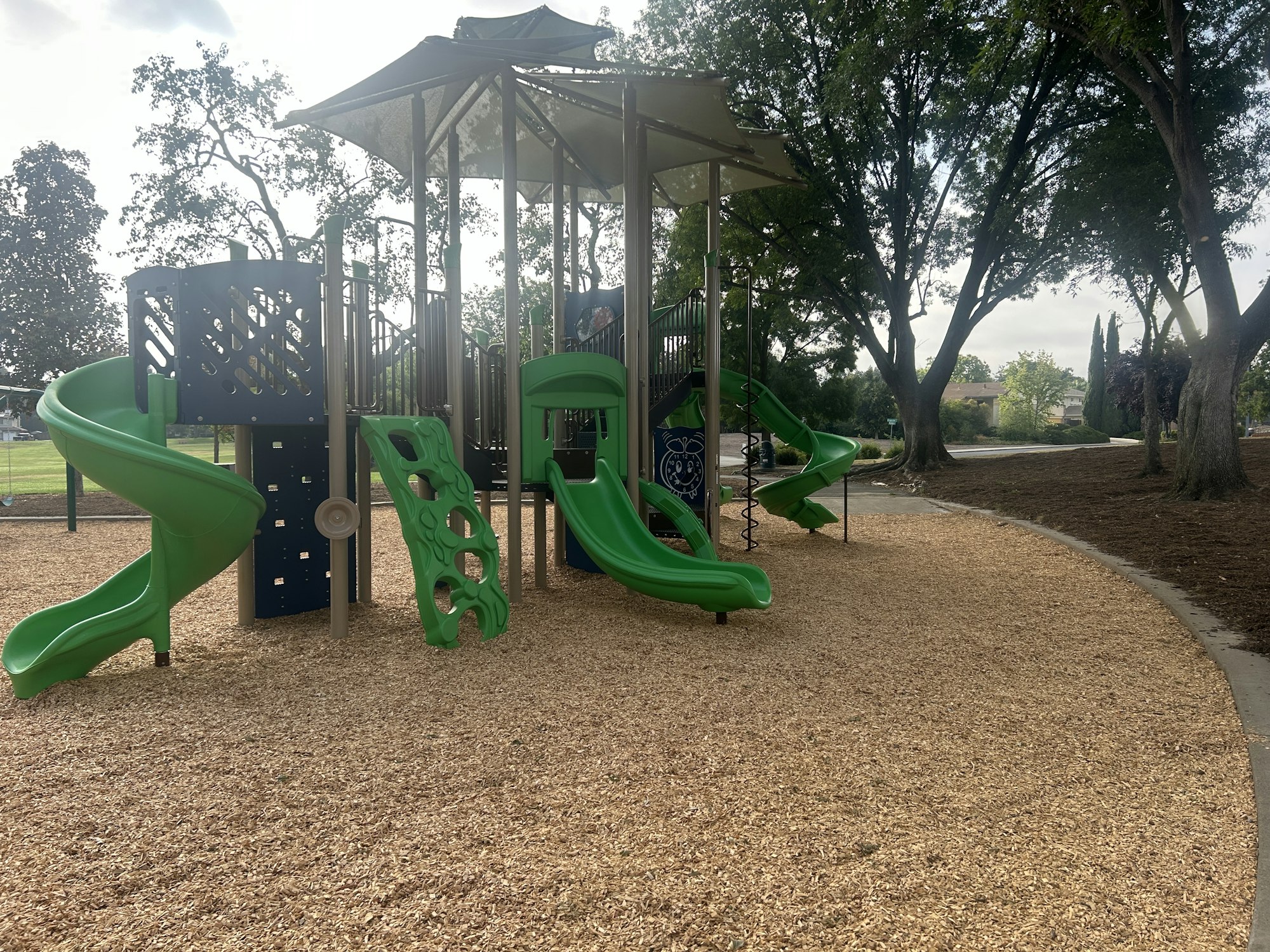 Playground with green slides, climbing structures, wood chip ground, surrounded by trees.