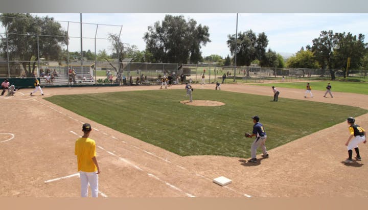 A youth baseball game in progress on a sunny day, with players on the field, a batter swinging, spectators in the stands.