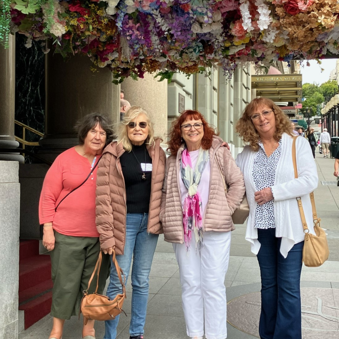 Four women posing under a colorful flower arrangement with a city street in the background.