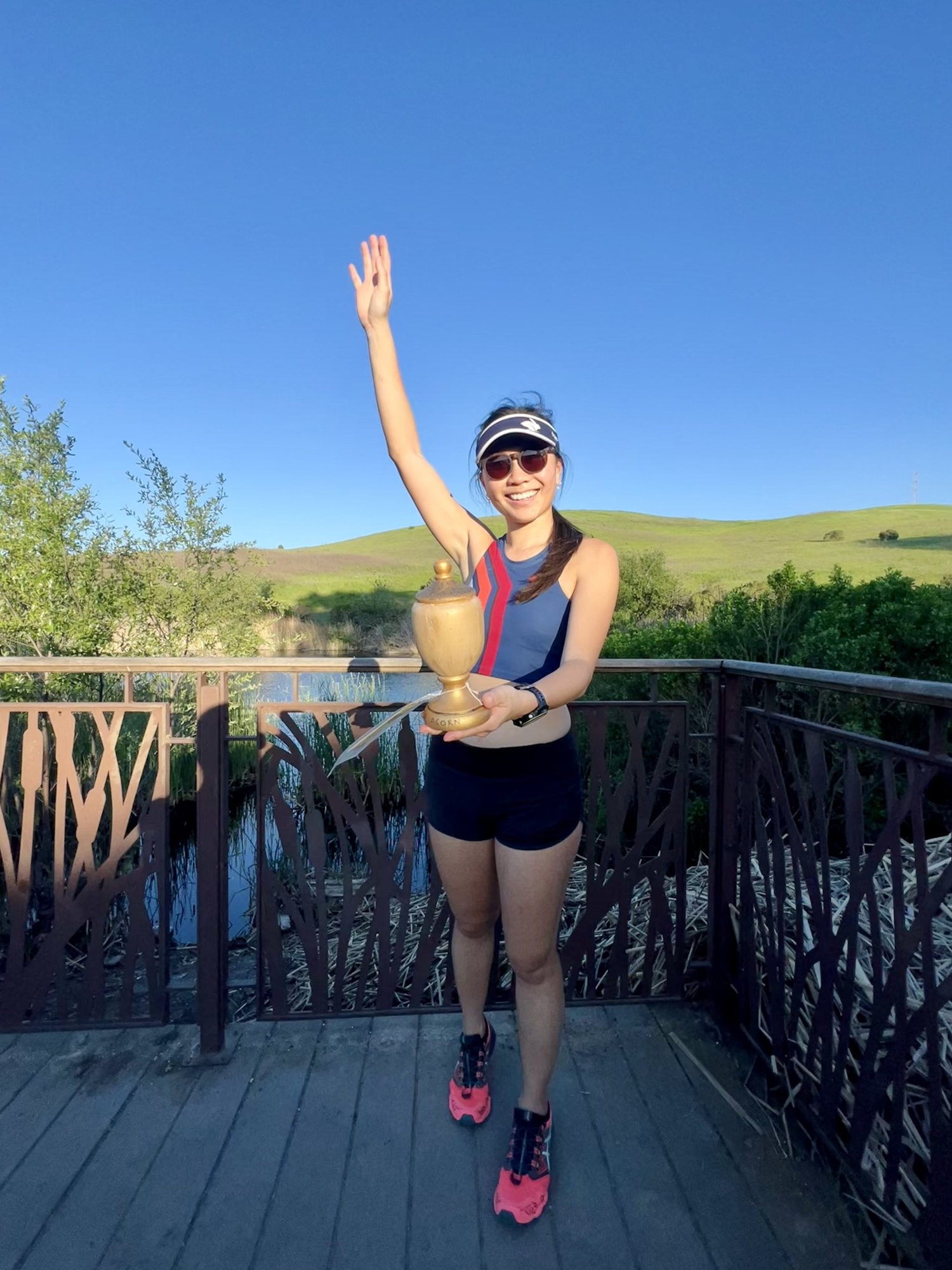 A happy woman with a trophy, wearing sportswear, on a bridge, sunny day with hills in the background.