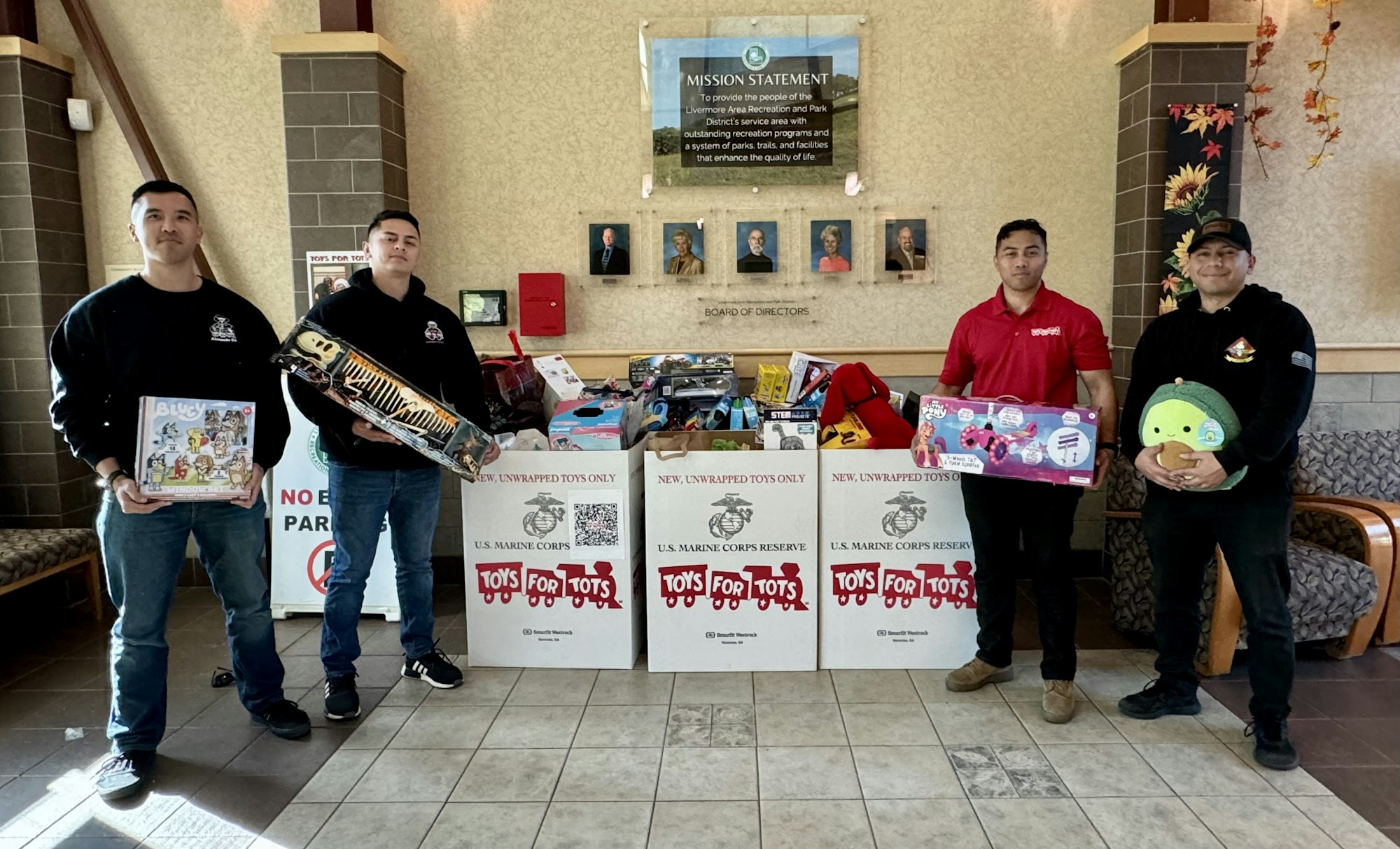 Four people holding toys stand by "Toys for Tots" bins, filled with toys for donation in a decorated lobby.
