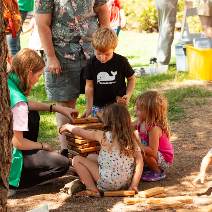 Children and an adult are playing outdoors, stacking large wooden blocks on the ground.