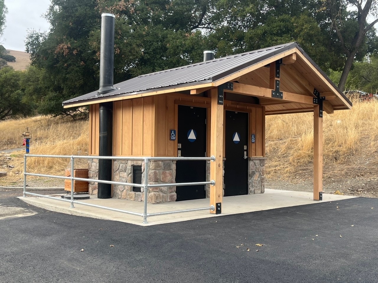 A small public restroom building with two doors, accessible signs, and a metal railing, set in a natural outdoor environment.