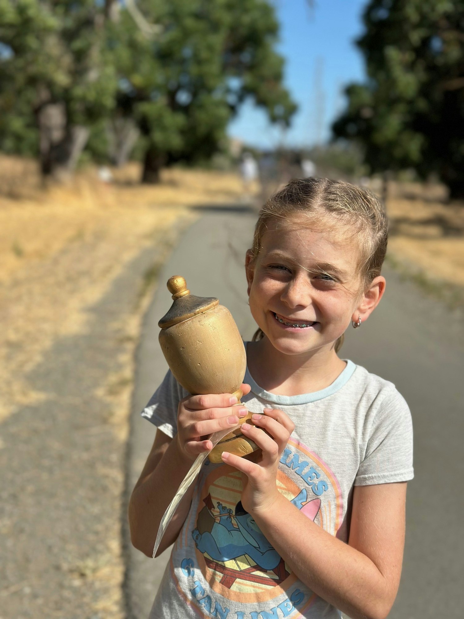 A smiling child outdoors holding a golden trophy on a sunny day.
