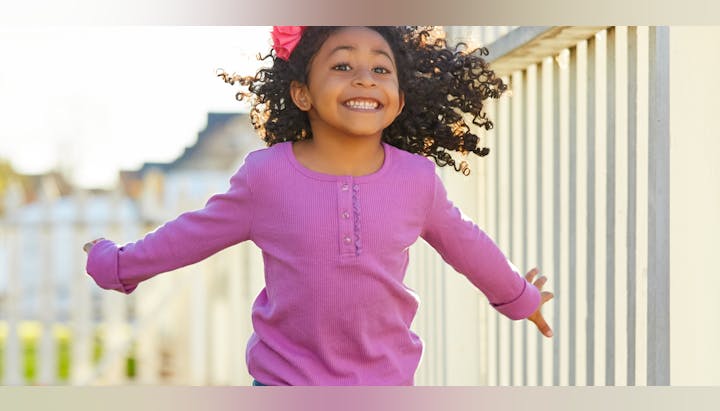 A joyful child in a pink shirt with curly hair jumps while smiling widely, outdoors near a fence.