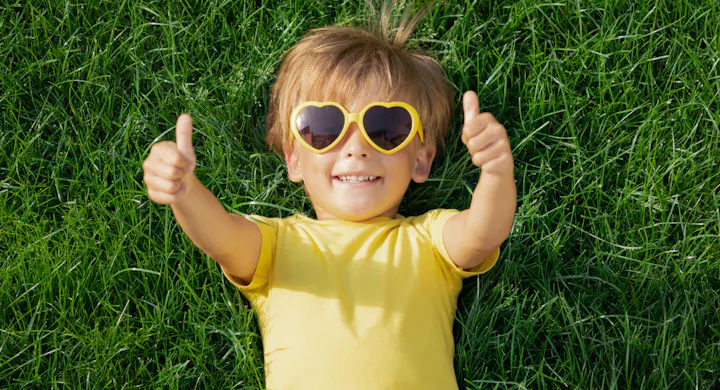 A cheerful child wearing heart-shaped sunglasses lies on grass, giving thumbs up while smiling in a sunny setting.