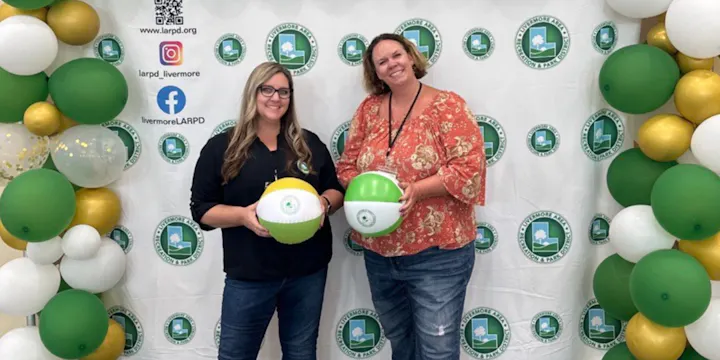 Two people holding beach balls in front of a Livermore Park District backdrop with balloons around them.