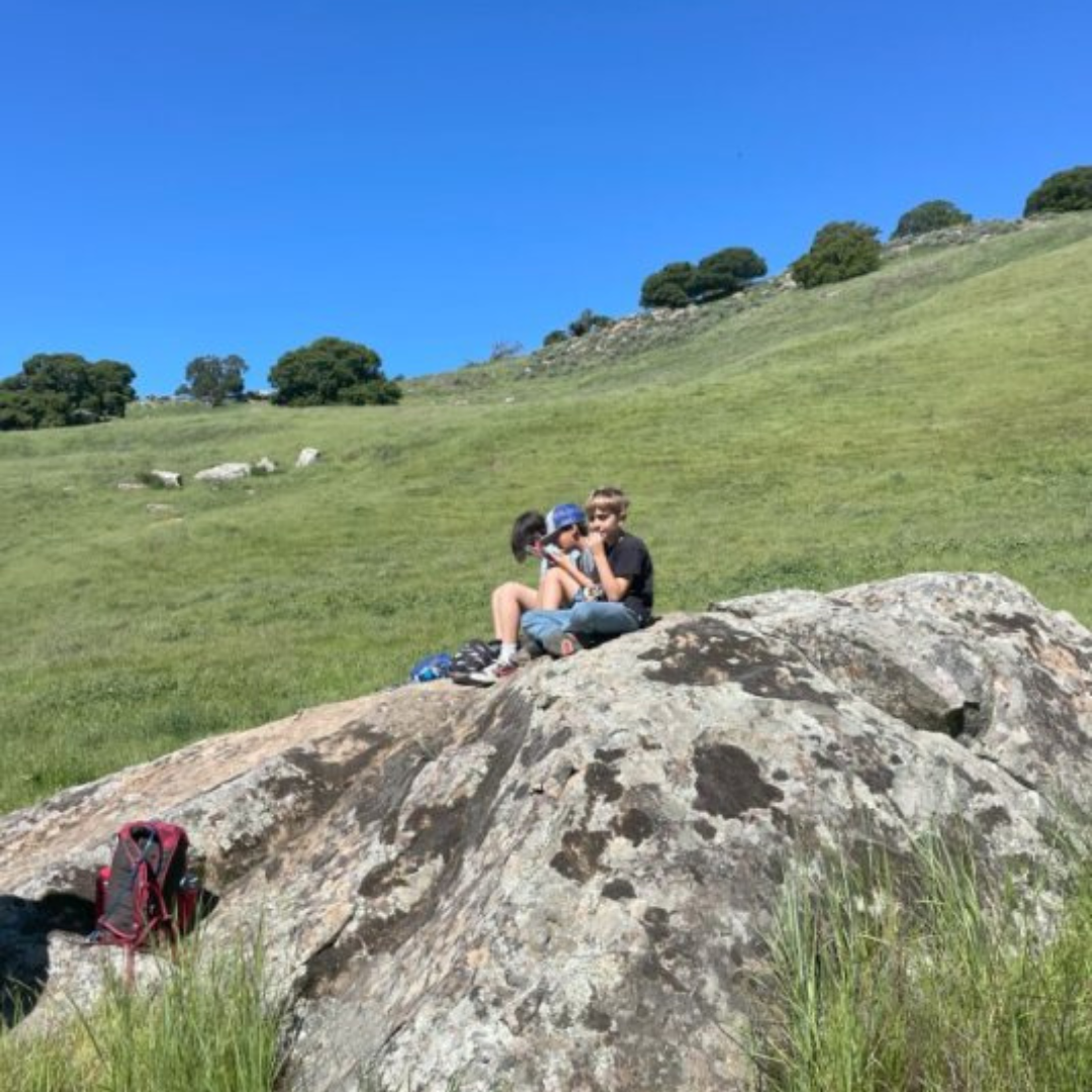 Three people on a rock in green grassland, with clear skies and a backpack nearby.