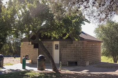 A small brick building with a tree overhead, trash can, and bench in a park setting.