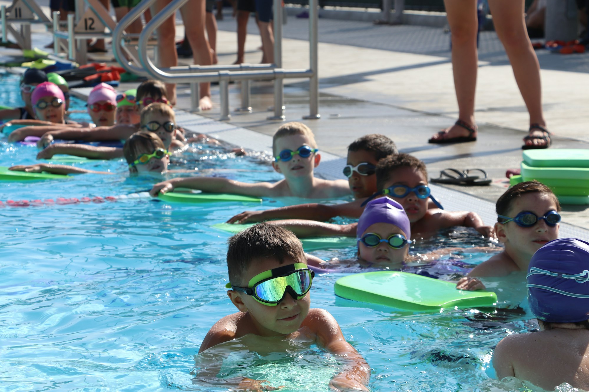 Kids in a swimming pool holding kickboards, wearing goggles and swim caps, lined up by the pool edge.