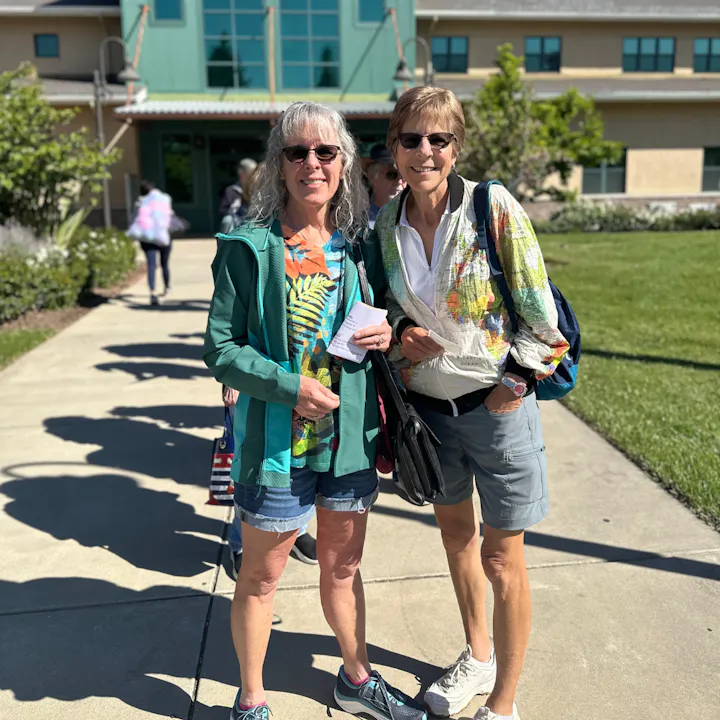 Two women smiling outdoors, dressed casually, standing on a walkway with greenery and a building in the background.