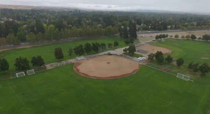 Aerial view of a sports field with soccer fields and a baseball diamond, surrounded by trees and a road.
