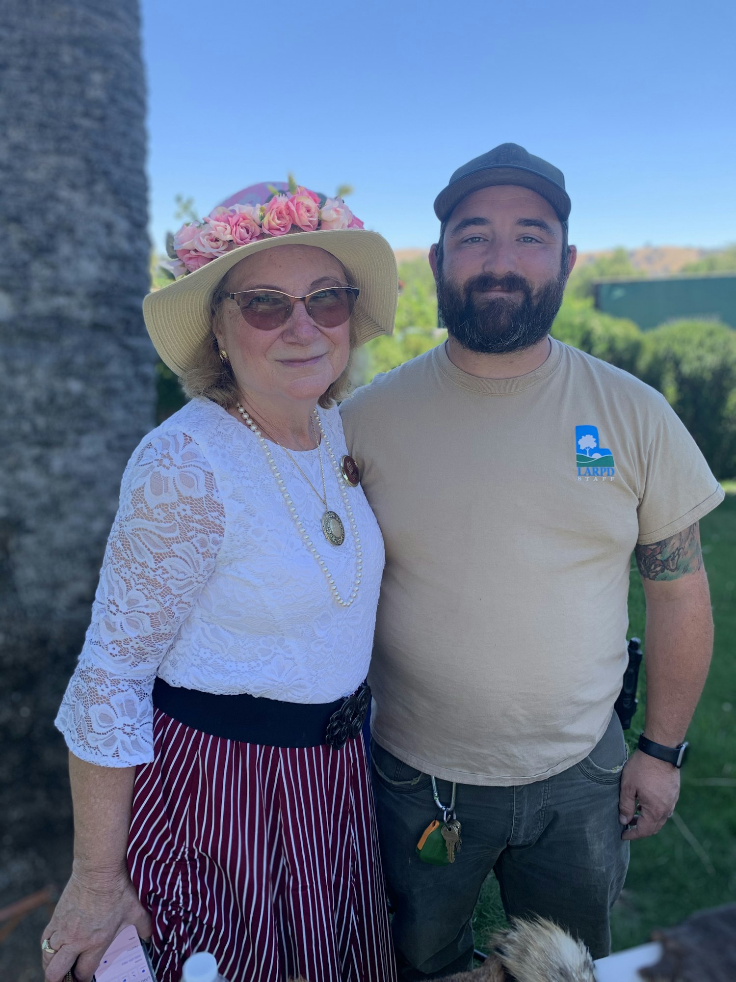 A woman in a flower hat and a man in a shirt pose together outdoors, smiling against a sunny background.