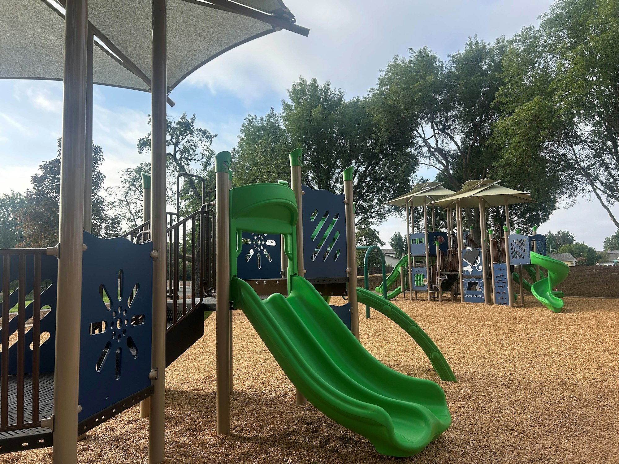 Playground with slides, climbing structures, and wood chips, surrounded by trees.