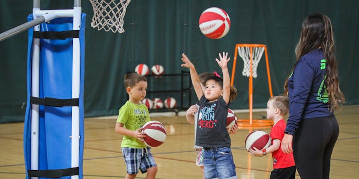 Children practicing basketball with an adult on a court, holding red and white basketballs.