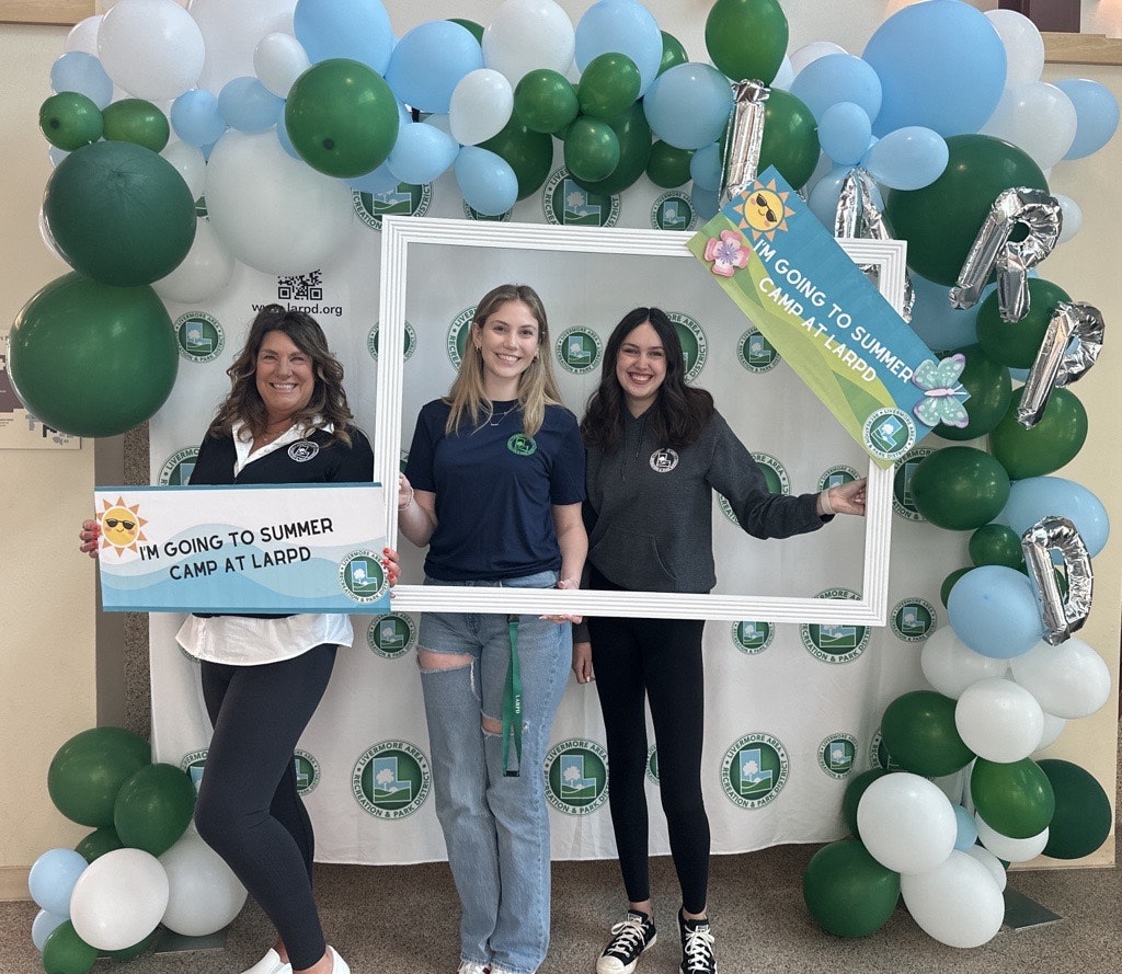 Three women pose with signs promoting summer camp at LARPD, surrounded by green and blue balloons in a festive setting.