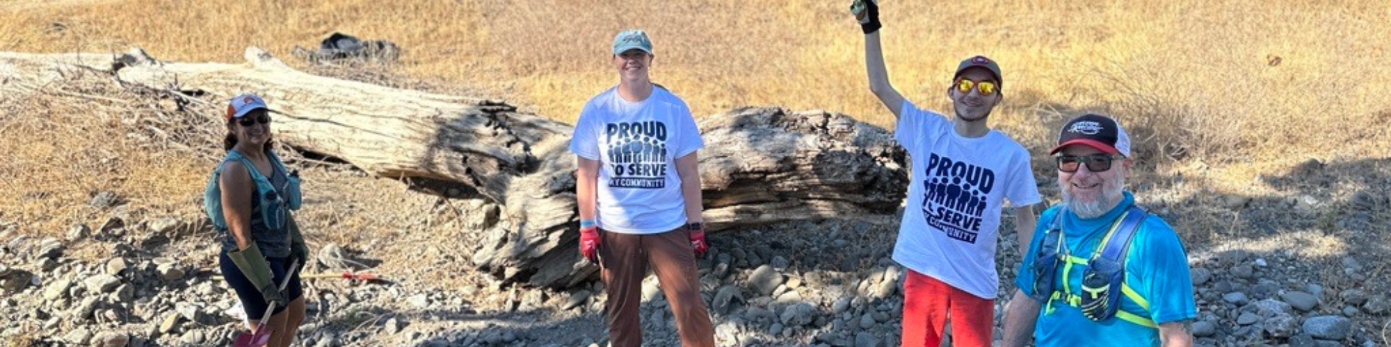 Four people outdoors in a dry area with a fallen tree. Some wear "Proud to Serve" shirts, suggesting a community event or cleanup.