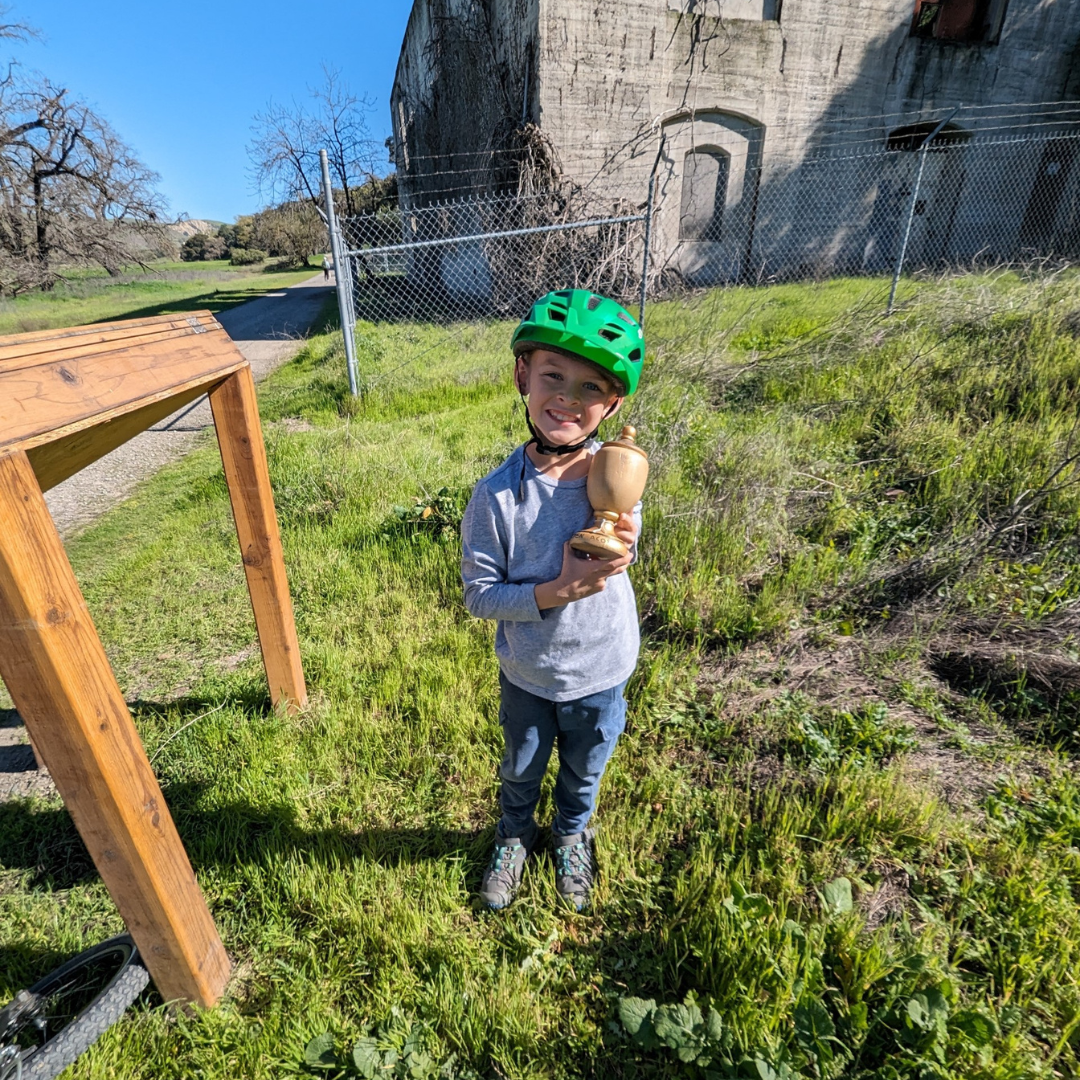 A smiling child wearing a green helmet, holding a wooden model, with a grassy backdrop and a concrete structure behind a fence.