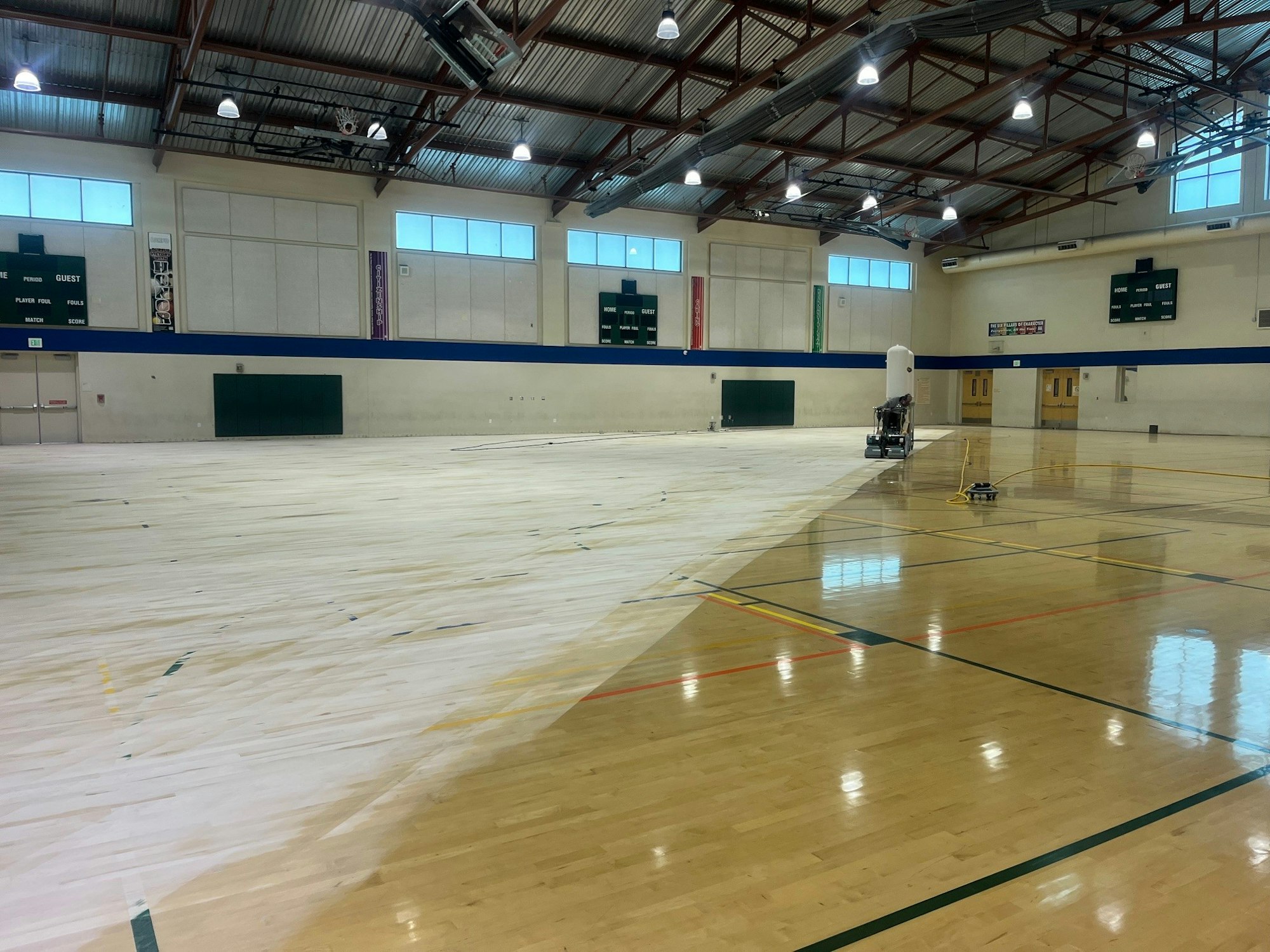 An indoor gymnasium with a partially resurfaced wooden floor and maintenance equipment.