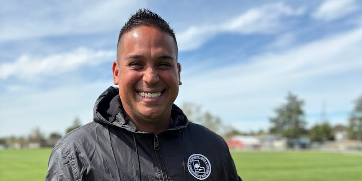 A smiling person in a jacket stands on a grassy field under a clear sky, conveying a positive and cheerful vibe.