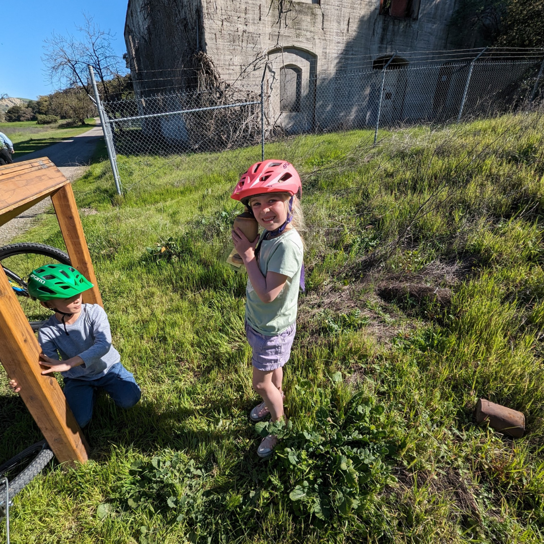 Two children with bike helmets outdoors, one standing and smiling, the other sitting; grassy area and concrete structure in background.