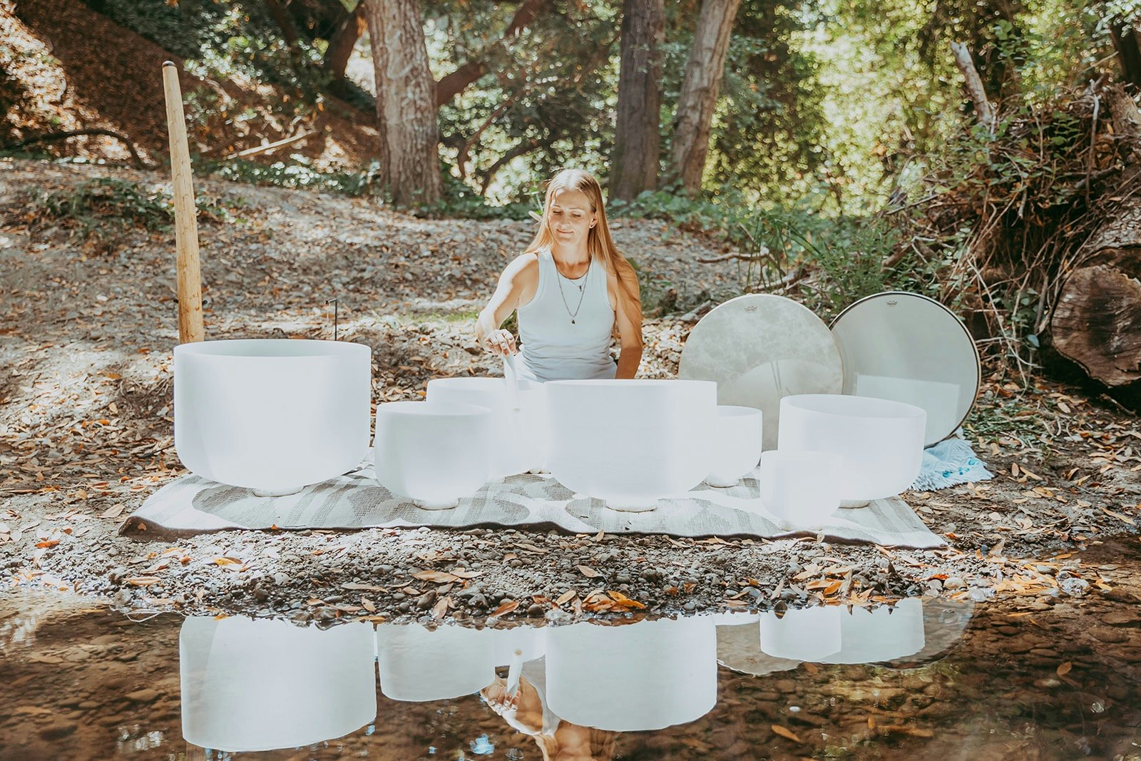 A person plays crystal singing bowls in a wooded area by the water.