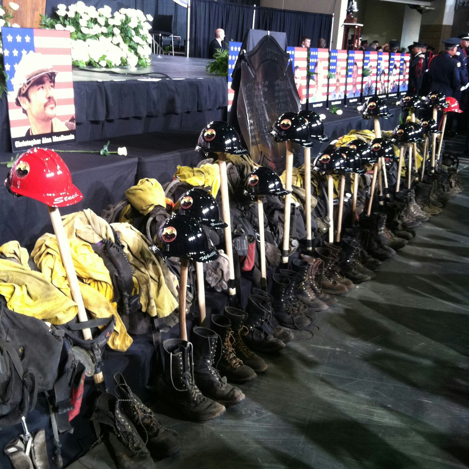 A memorial with firefighter helmets, boots, and jackets lined up in front of a stage with flowers and US flags.