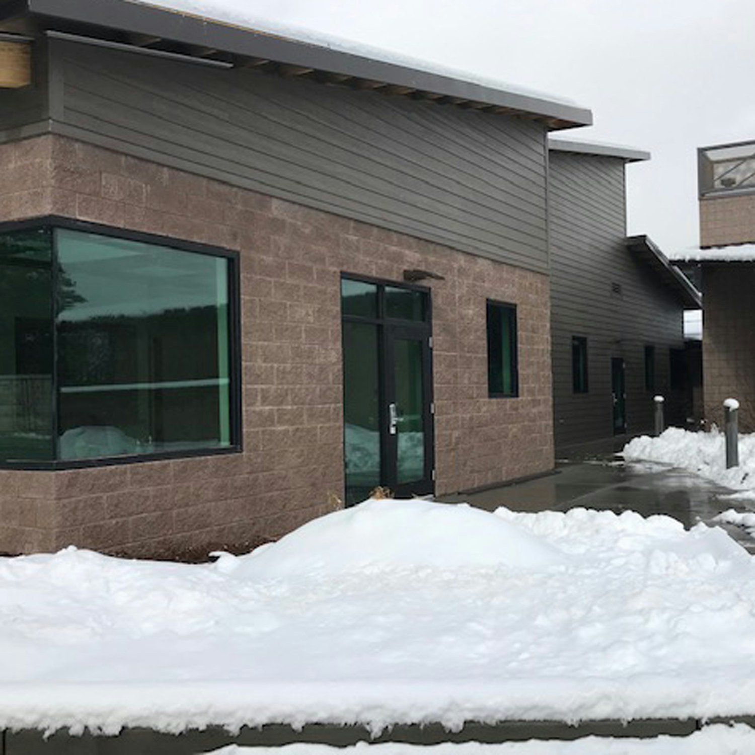 A modern building with large windows, partially covered sidewalk, and snow piled up in front.