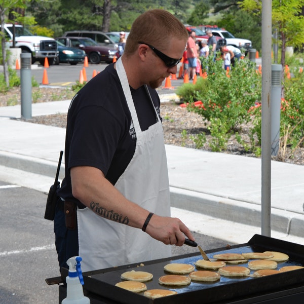 A man in an apron and sunglasses cooks pancakes on a griddle outdoors with onlookers in the background.