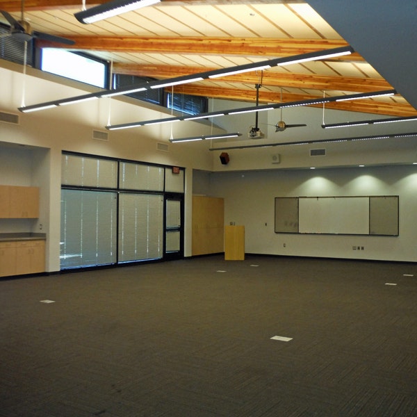 An empty modern classroom with a whiteboard, projector, kitchenette, and large windows allowing natural light.