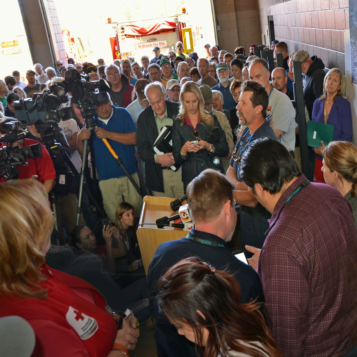 A crowded press conference with reporters, cameras, and onlookers in a garage-like setting.