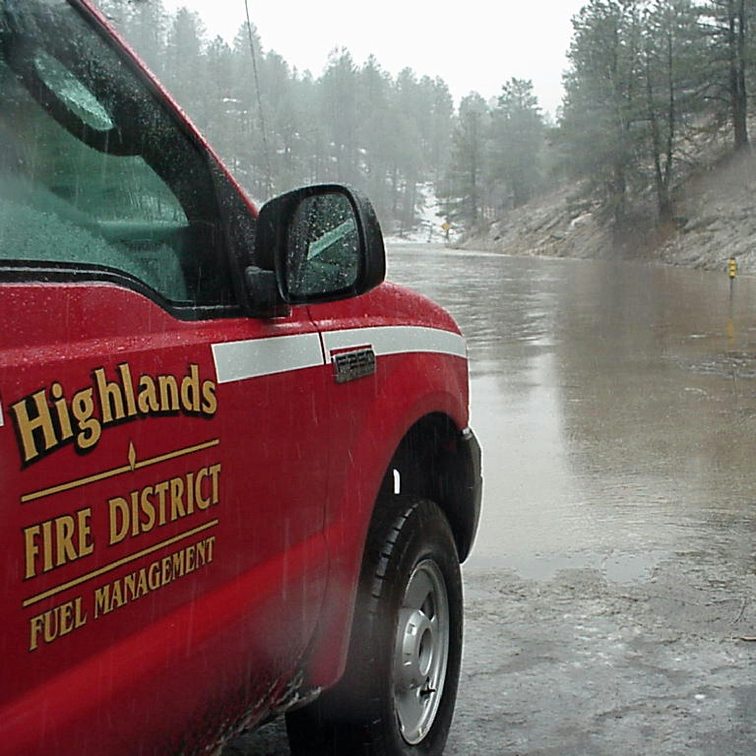 A red vehicle with "Highlands Fire District" text, parked on a rain-soaked road surrounded by trees.