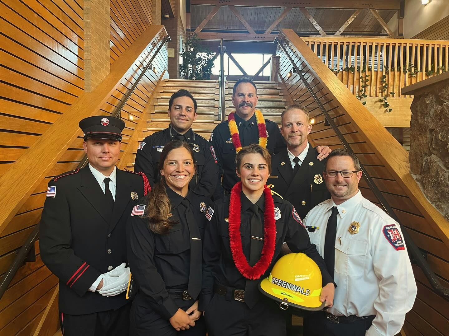 A group of seven smiling firefighters in uniform on a staircase, one holding a yellow helmet.