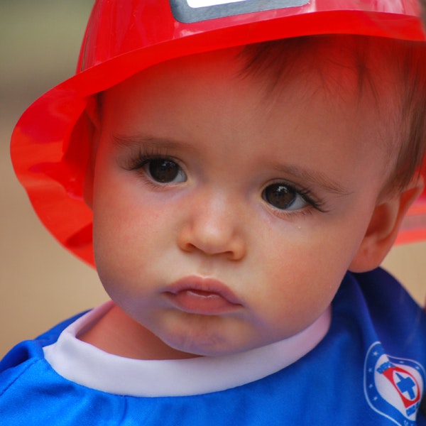 A toddler wearing a red firefighter hat and a blue outfit with a badge.