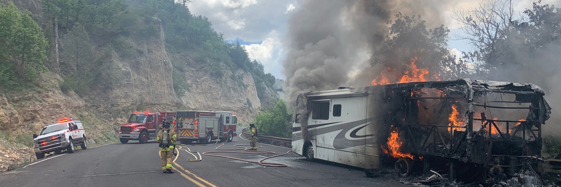 Firefighters tackling a burning RV on a road near mountainous terrain.