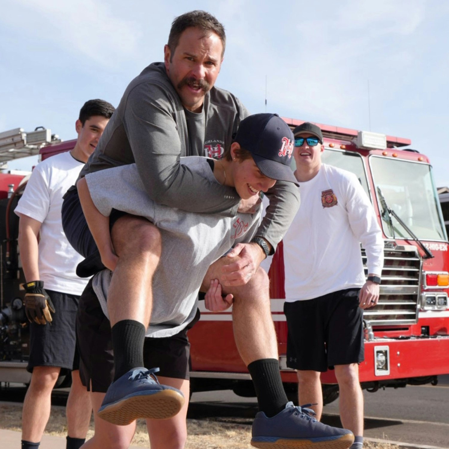 A person gives another a piggyback ride in front of a fire truck, with two others nearby smiling and watching.