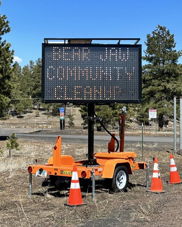 Portable electronic sign by road displaying "BEAR JAW COMMUNITY CLEANUP" with orange traffic cones around.