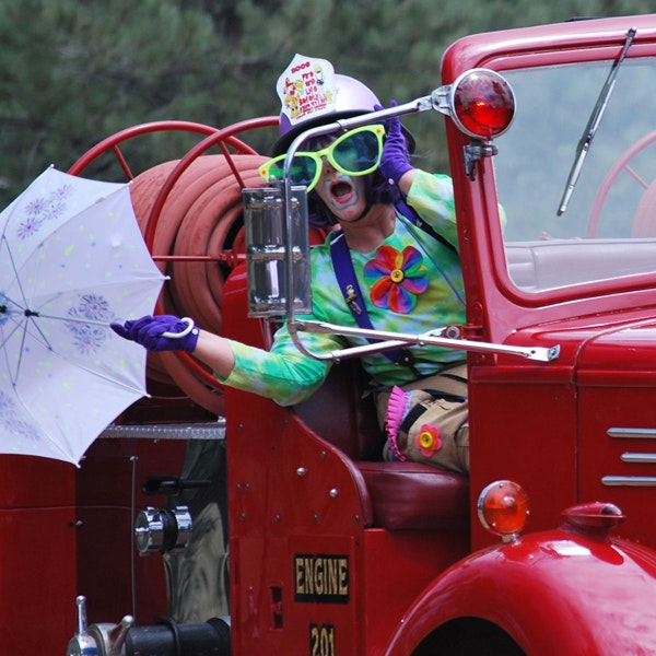 Person in colorful clown outfit with goggles, driving a vintage fire engine, holding an umbrella. #Clown #FireTruck #Fun