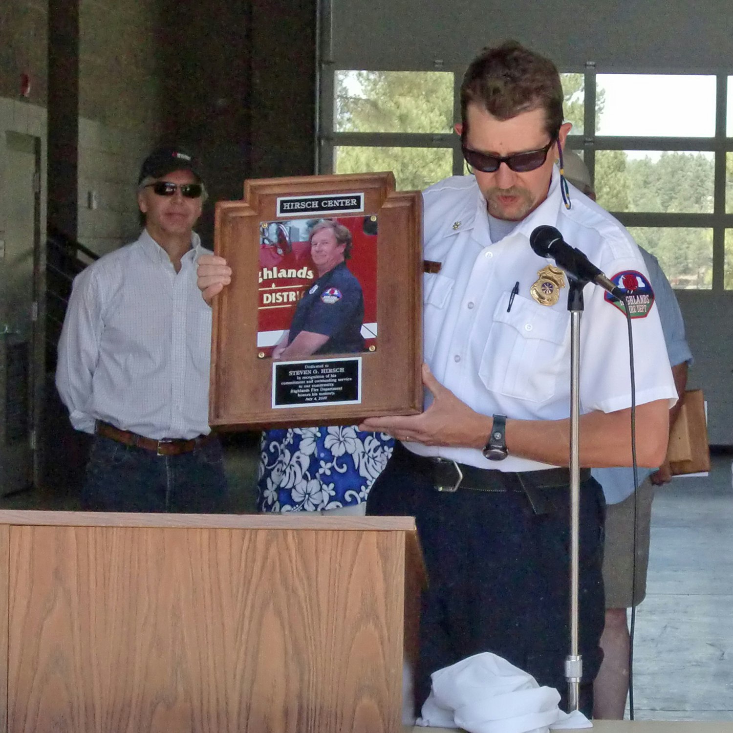 A man in a white shirt presents a commemorative plaque while another looks on, in an outdoor event with a microphone stand.