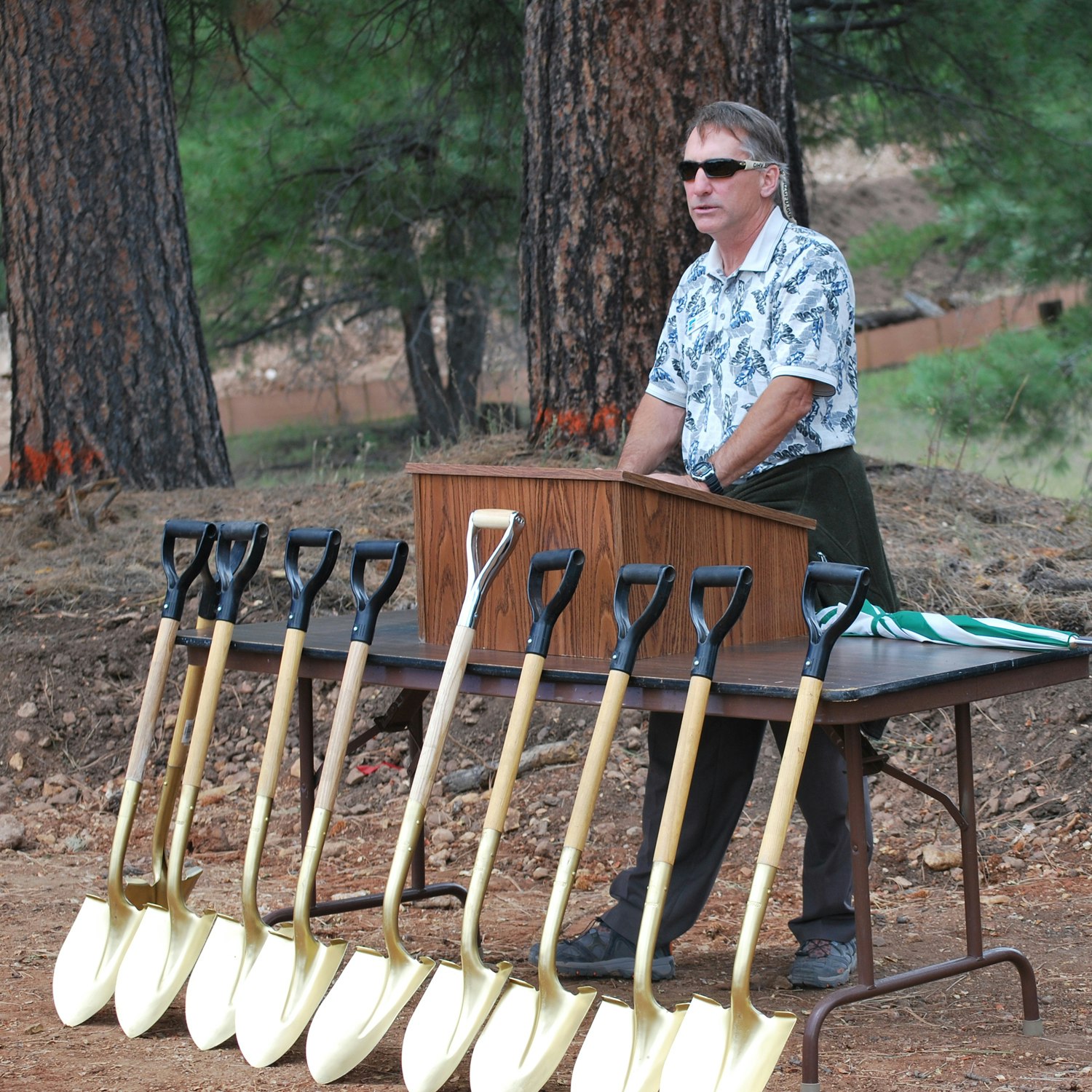 Man standing behind table with ceremonial shovels, likely at a groundbreaking event. Trees in the background.