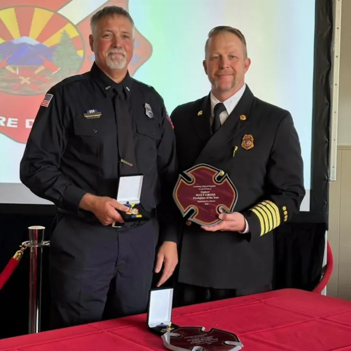 Two firefighters are posing together, one holding a medal and the other a plaque recognizing him as "Firefighter of the Year."