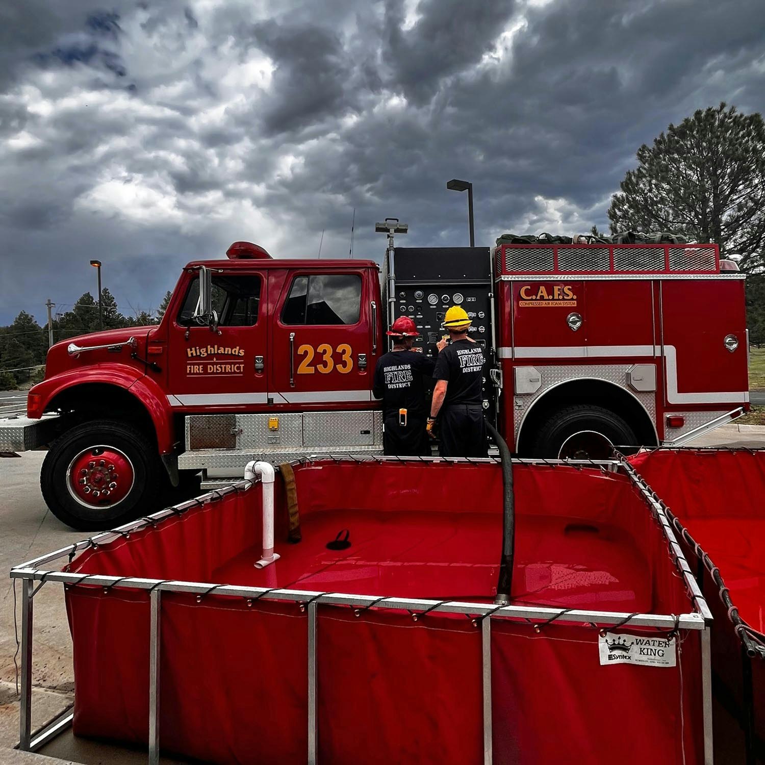 A fire truck and firefighters beside a temporary water tank under a cloudy sky.