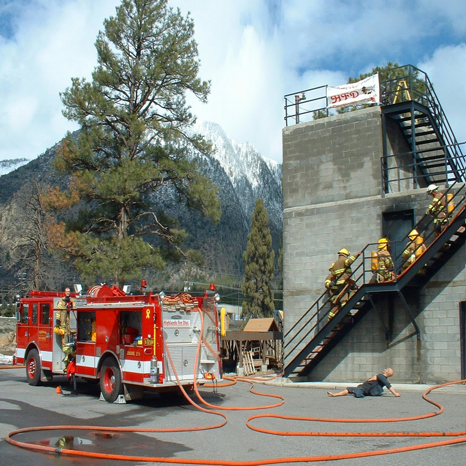 Fire truck at a training exercise with firefighters on stairs and a dummy on the ground, mountains and trees in the background.